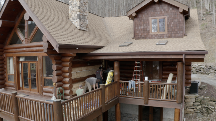 Log cabin home undergoing restoration work with workers on the deck replacing logs, featuring stone and wood construction with tan roof and wooden railings, situated on a hillside in a wooded area